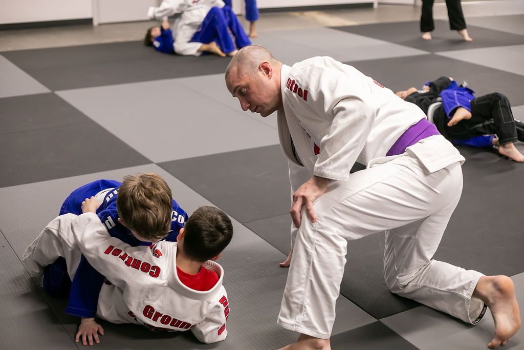 Two martial arts students grapple on mats while an instructor in a white gi watches nearby.