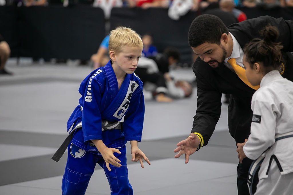 Two kids in blue and white gis practice grappling on a mat with a coach nearby