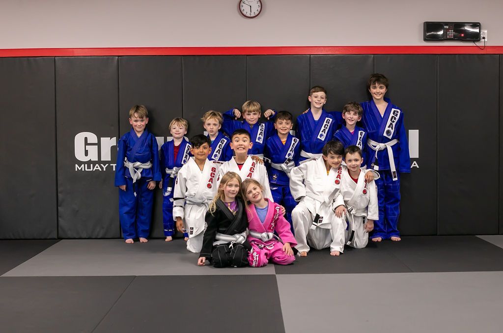 Group of martial arts students in blue, white, and pink uniforms posing on mats in a gym.
