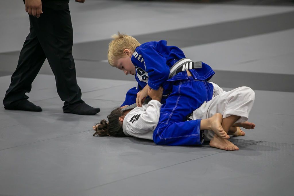 Two children in blue and white judo uniforms grappling on a mat while a referee stands nearby