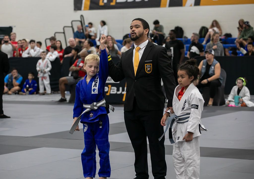 Martial arts competition with a referee standing between two children in blue and white gis on a gym mat