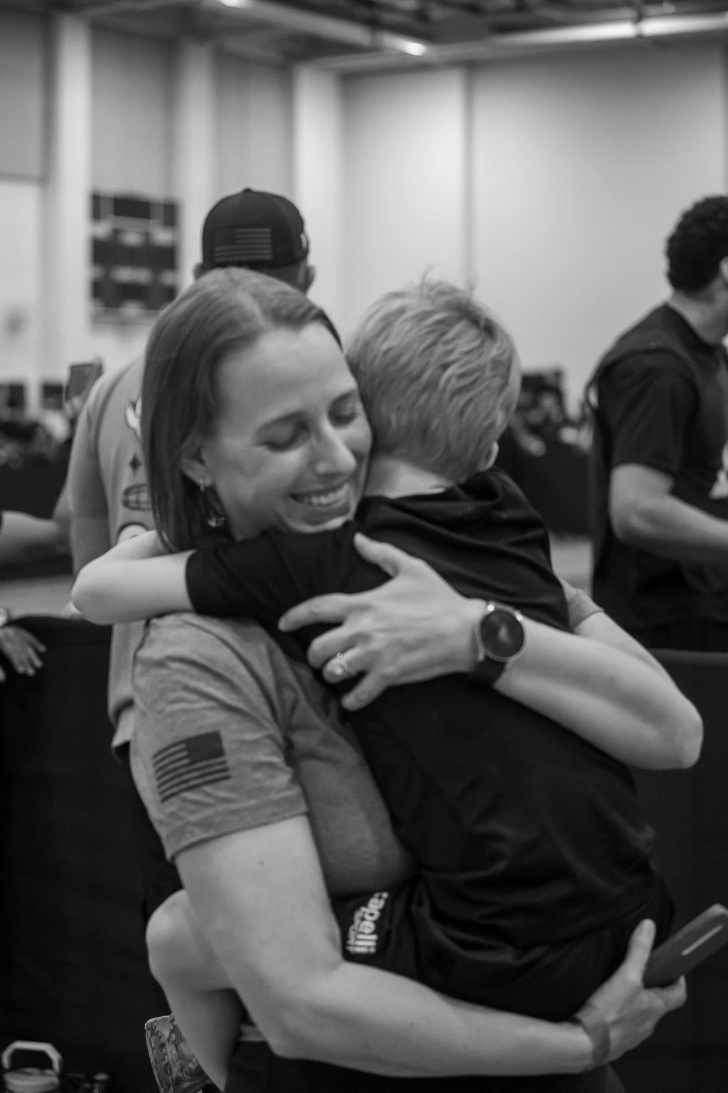 Woman hugging a child in a gym, both smiling, with other people in the background