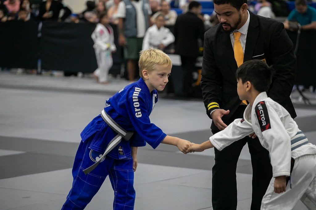 Two martial arts competitors in blue and white uniforms shake hands on a mat, with a referee nearby.