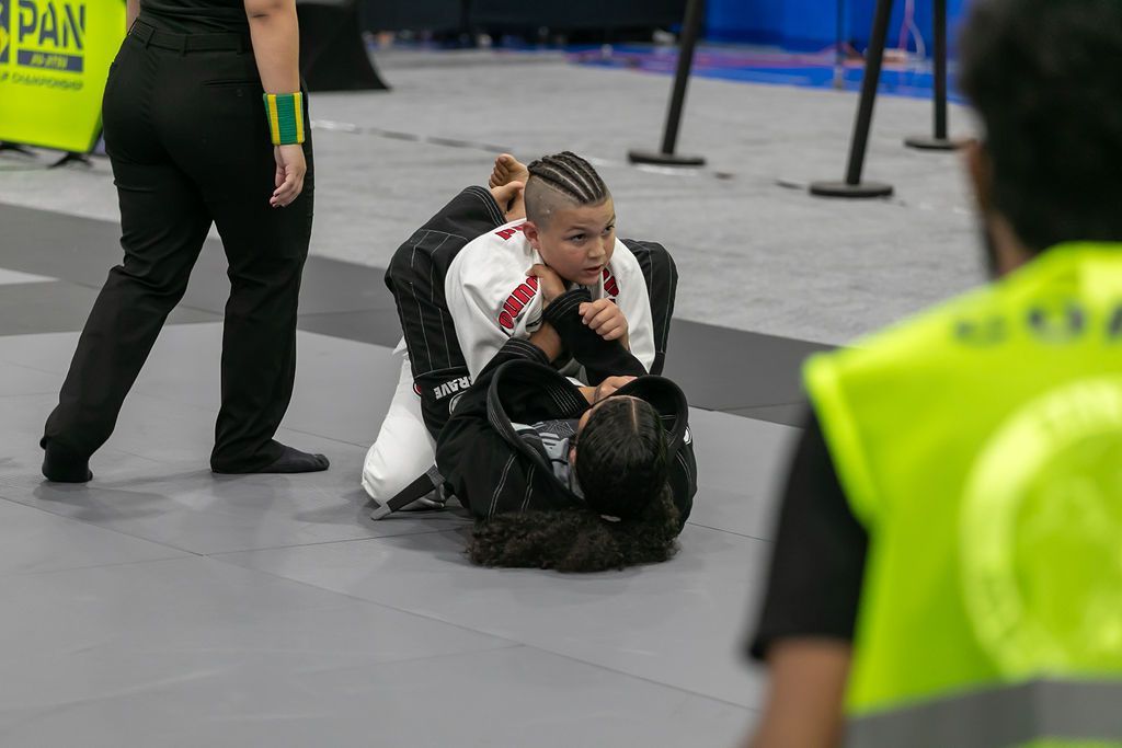 Two grapplers on a mat, one pinning the other during a match, with referees nearby