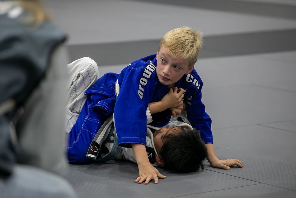 Two children practicing jiu-jitsu on a gray mat, one in a blue gi on top of the other.