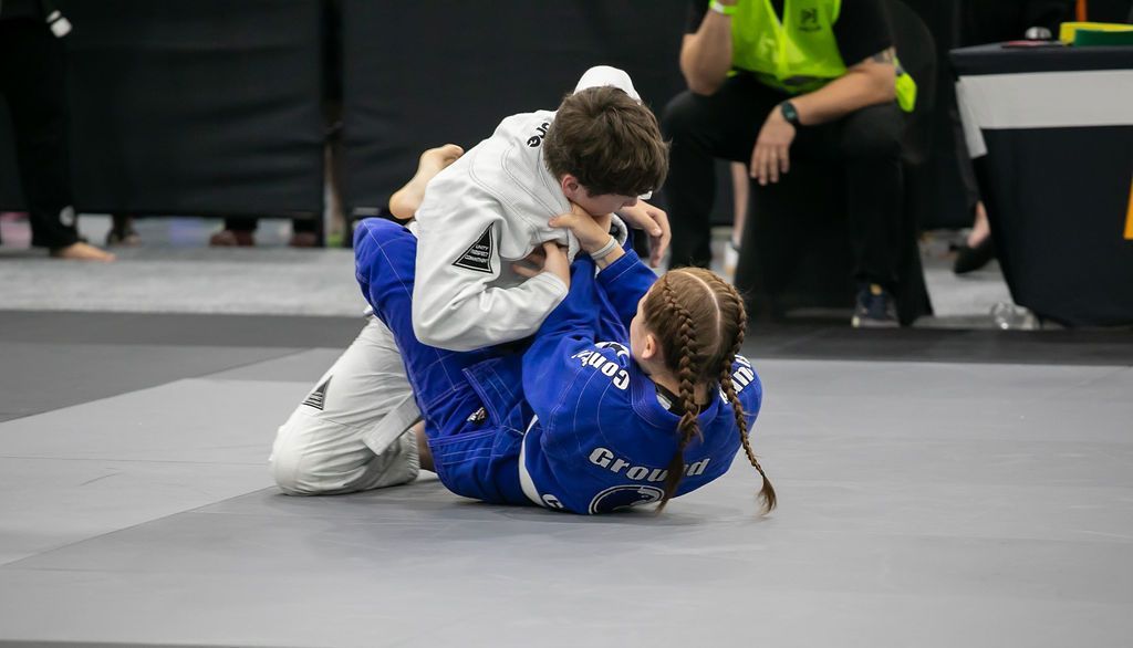 Two jiu-jitsu competitors grapple on a mat, one in blue on top of one in white.