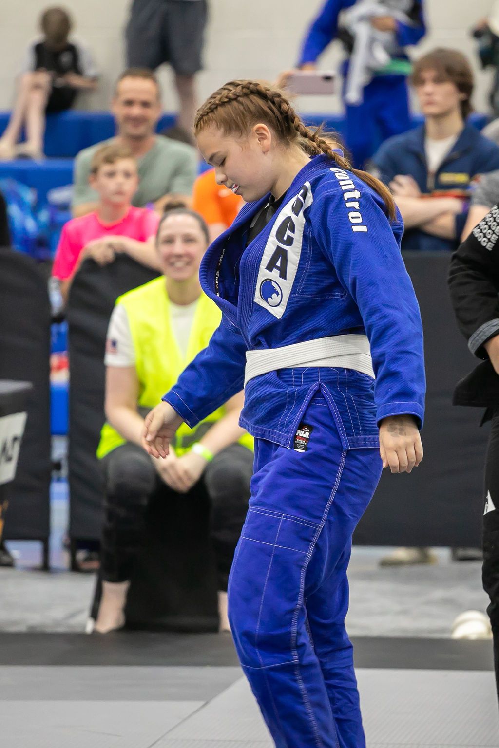Person in blue martial arts gi standing on a mat with spectators in the background