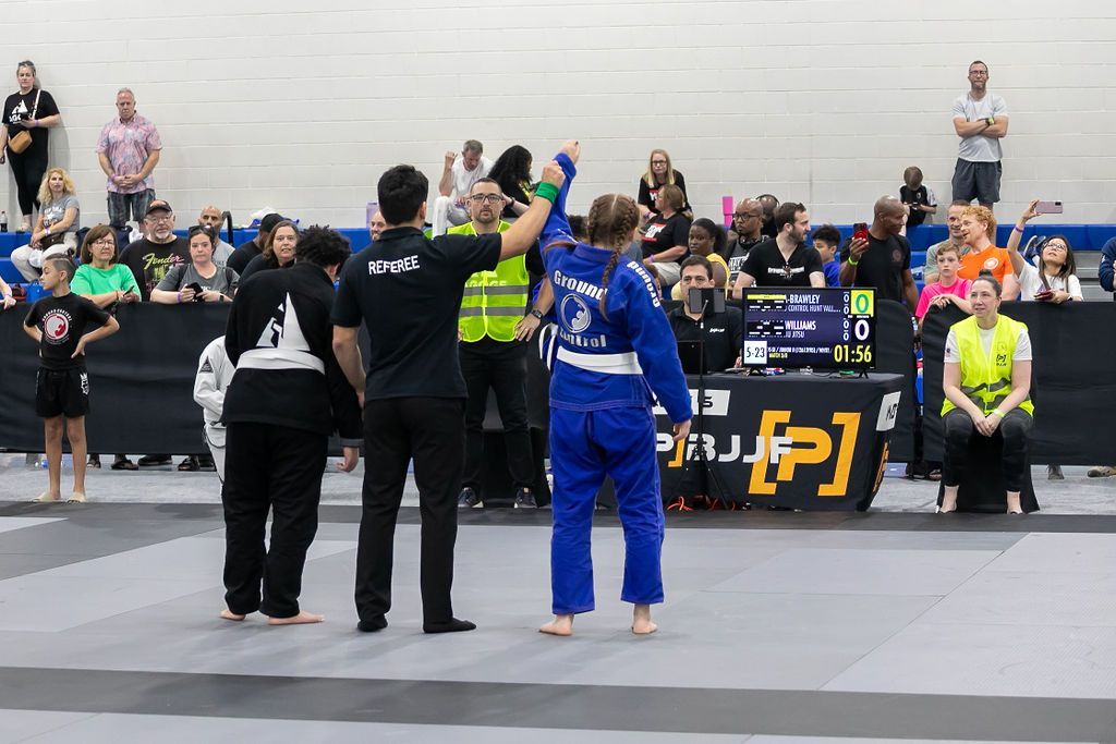 BJJ match in progress with two competitors and a referee on a mat, audience watching in the background