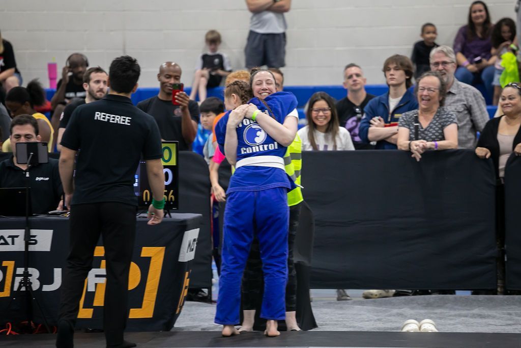 Two competitors in blue and black uniforms embrace on a mat before a crowd