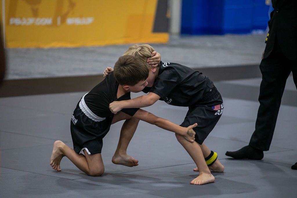 Two children grappling on a mat during a martial arts match, with a referee standing nearby.