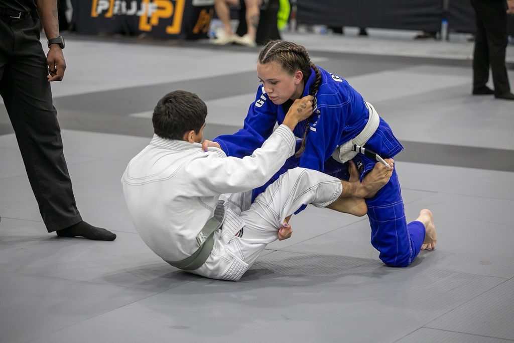 Two jiu-jitsu competitors grapple on a mat, one in a blue gi kneeling over a white-gi opponent.