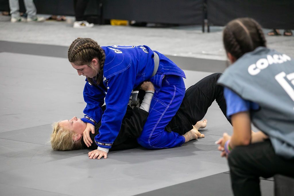 Two grapplers on a mat, one in a blue gi mounting the other during a jiu-jitsu match.