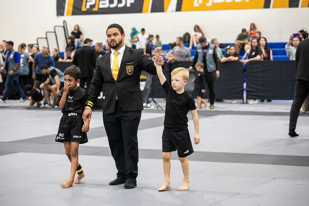 Two children in black uniforms stand beside a referee on a mat, with a crowd watching in the background.