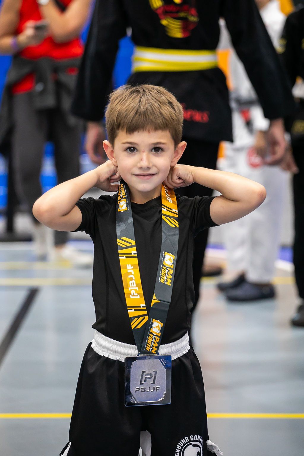 Young child in black outfit with gold medal, smiling and holding hands to ears at an indoor event.