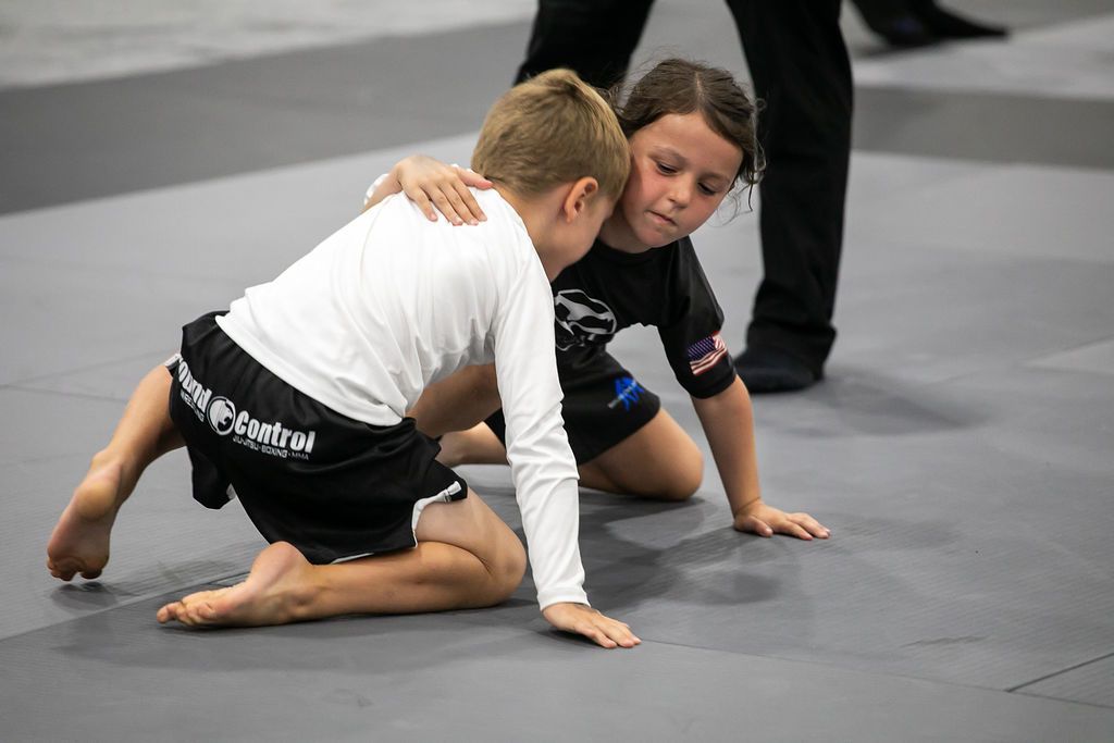 Two children grapple on a mat during a wrestling match, with a referee standing nearby.