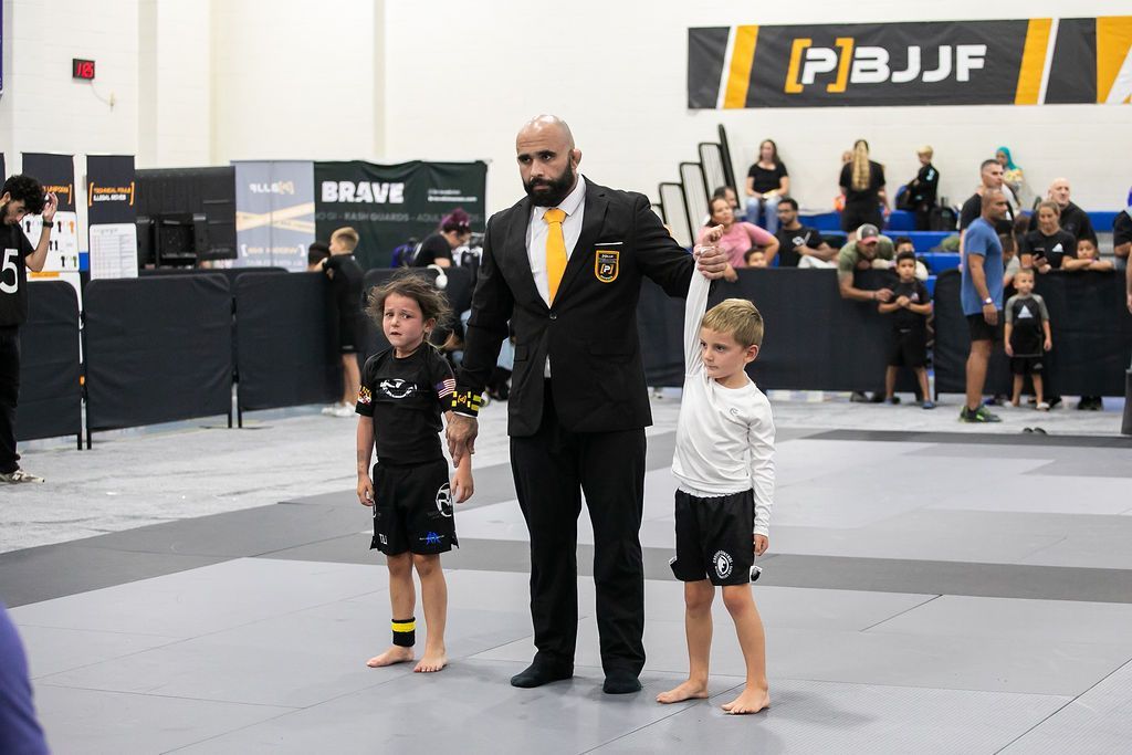 Referee standing between two young competitors on a jiu-jitsu mat at a tournament