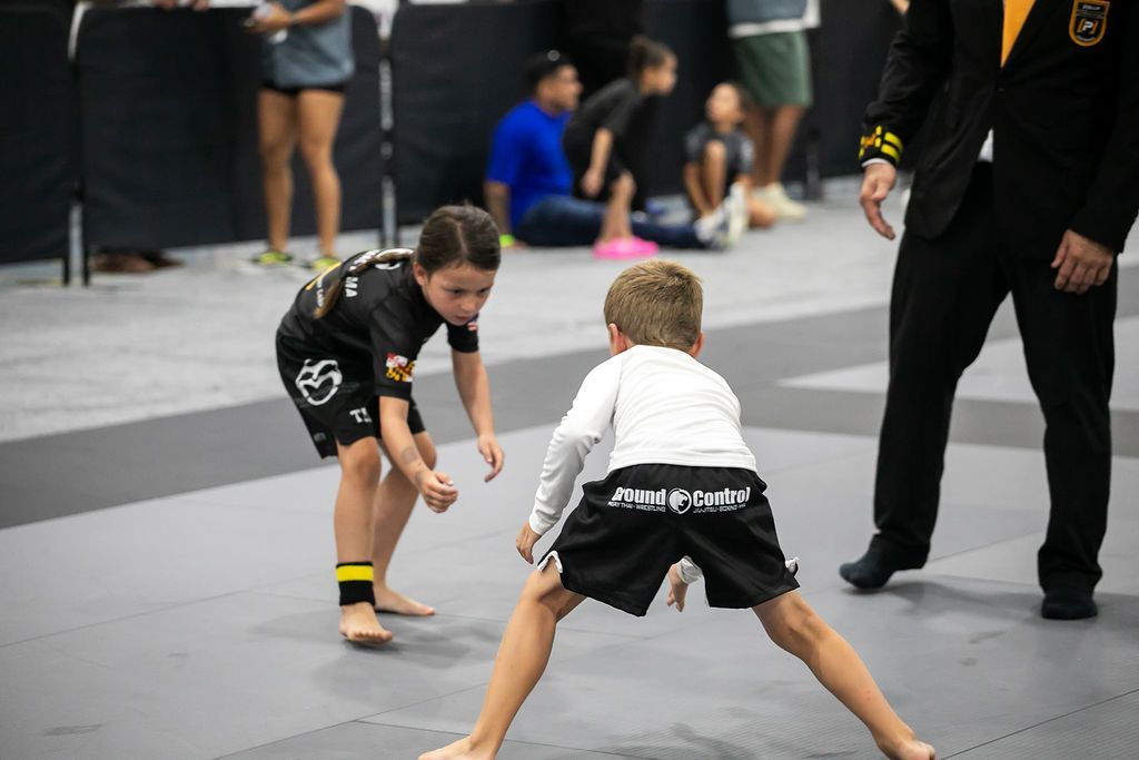 Two children in grappling stances on a mat during a martial arts match, with spectators nearby