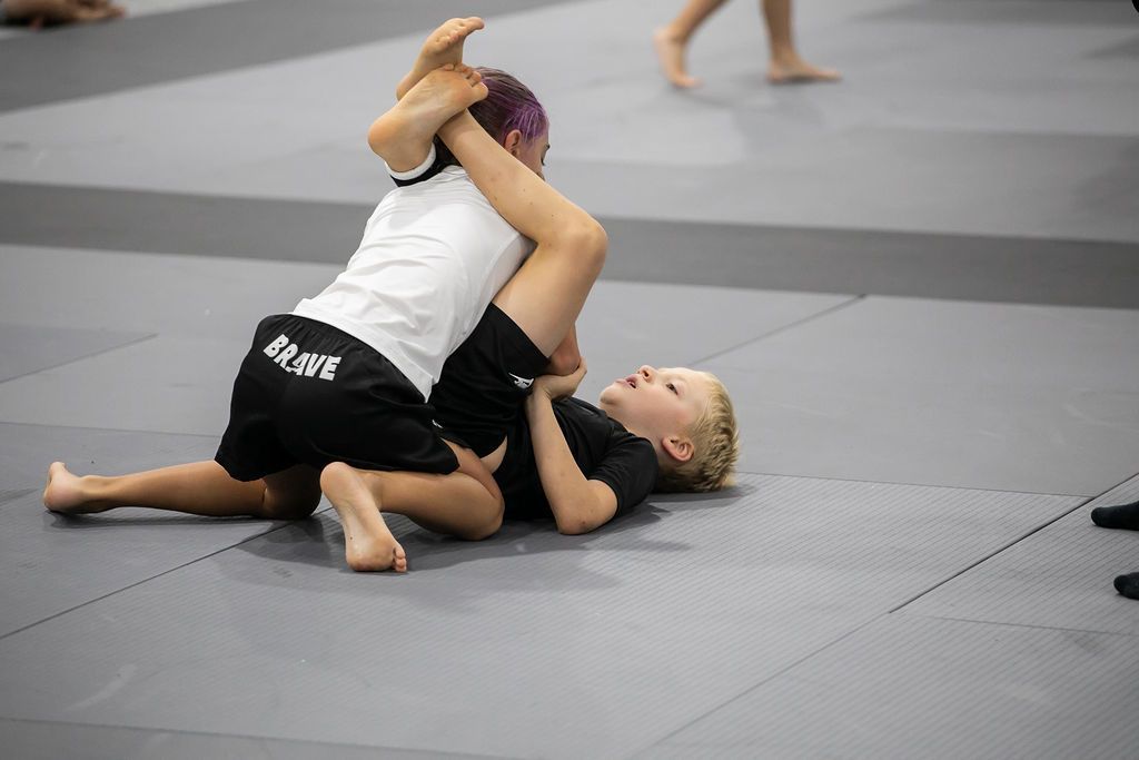 Two children wrestling on a gray mat in a gym, one in a white shirt on top.