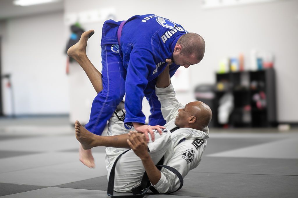 Two martial artists grappling on mats in a dojo, with two others blurred in the background.