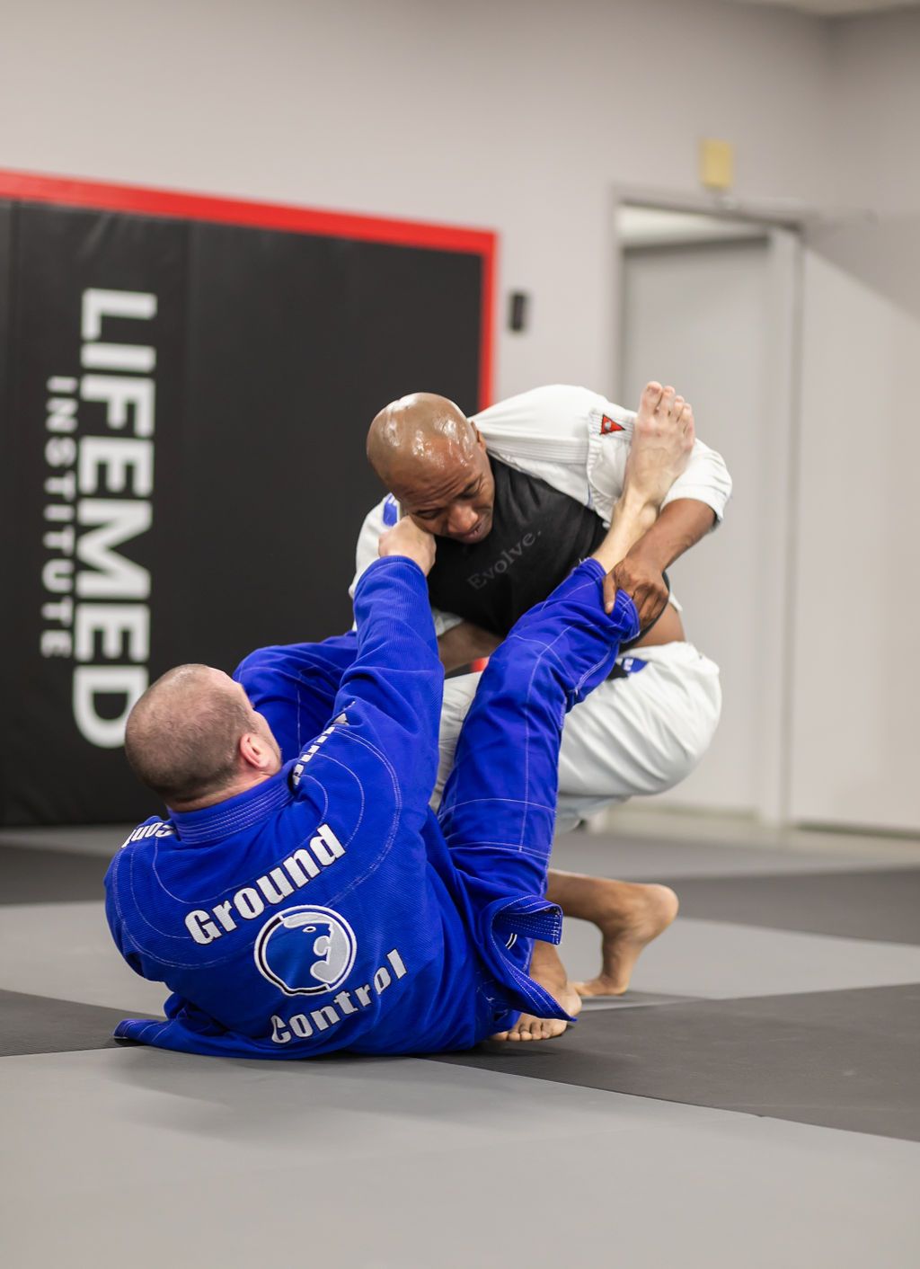 Two jiu-jitsu athletes grappling on mats, one in blue gi and one in white, inside a training gym.