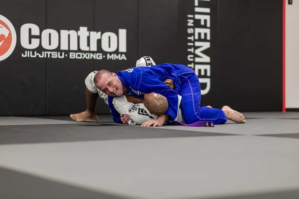 Two grapplers in blue and white gis on a mat, one in top control at a jiu-jitsu gym