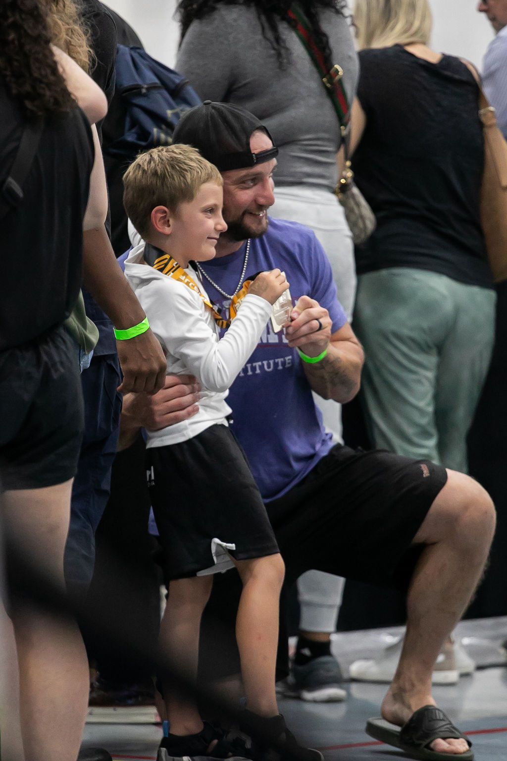 A man kneels beside a child in a white shirt at a crowded event, both clapping and smiling.