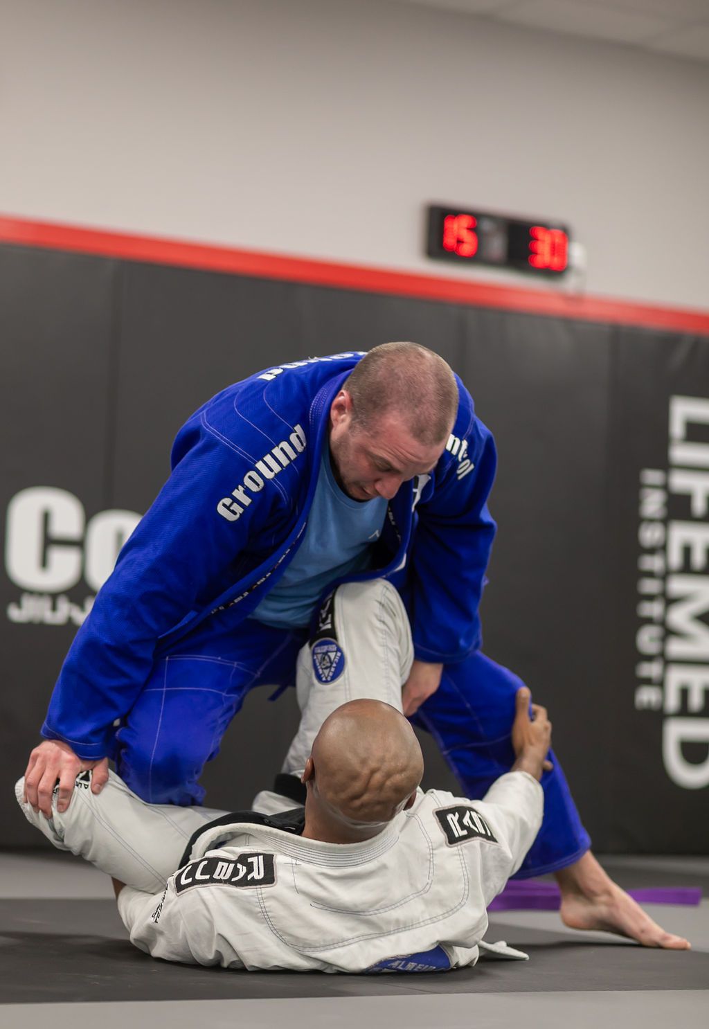 Two jiu-jitsu fighters grapple on a mat, one in a blue gi mounted over one in a white gi.