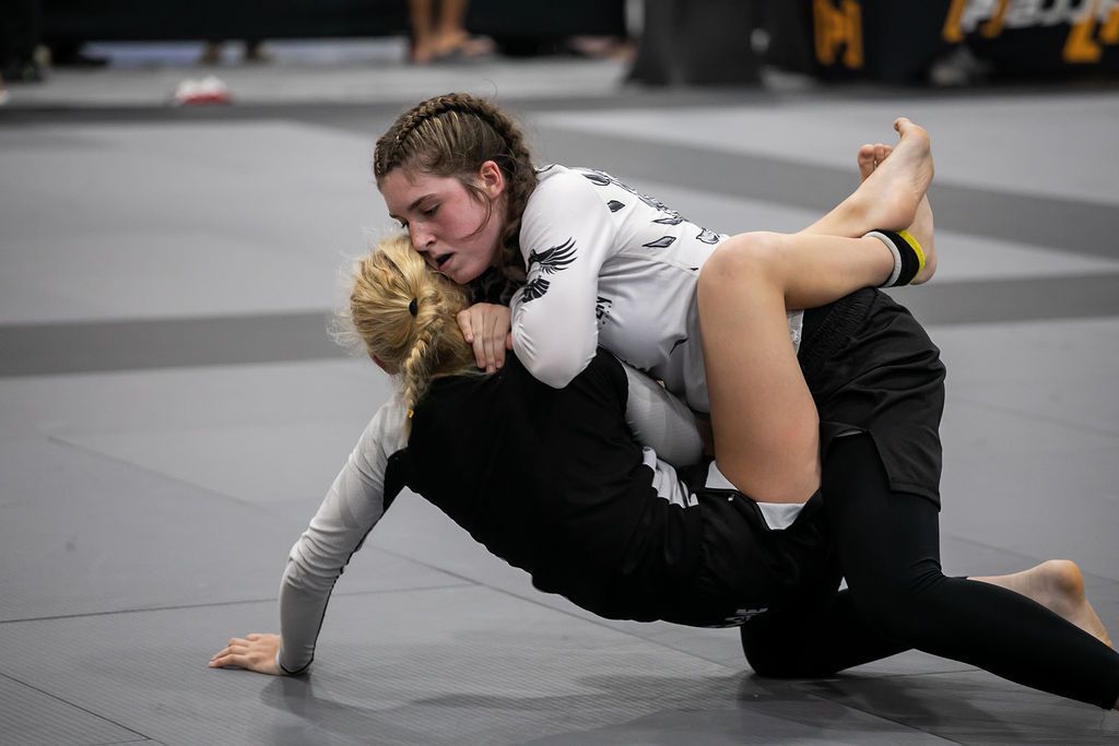 Two martial artists grappling on mats in a dojo, with two others blurred in the background.