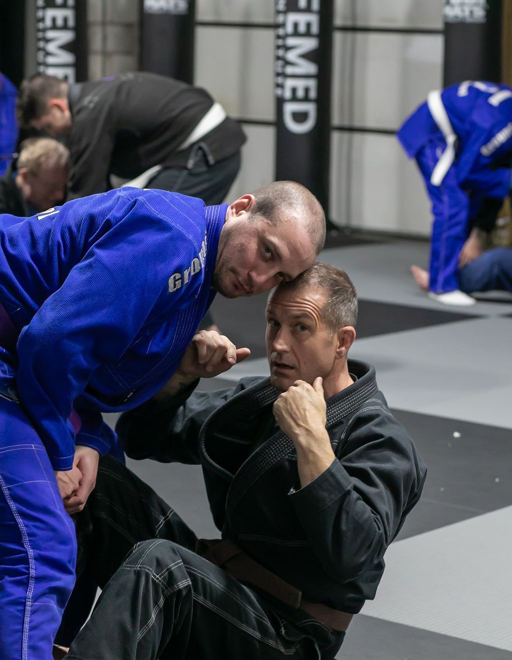 Two men in martial arts gis spar on mats, one leaning over the other in a training gym.