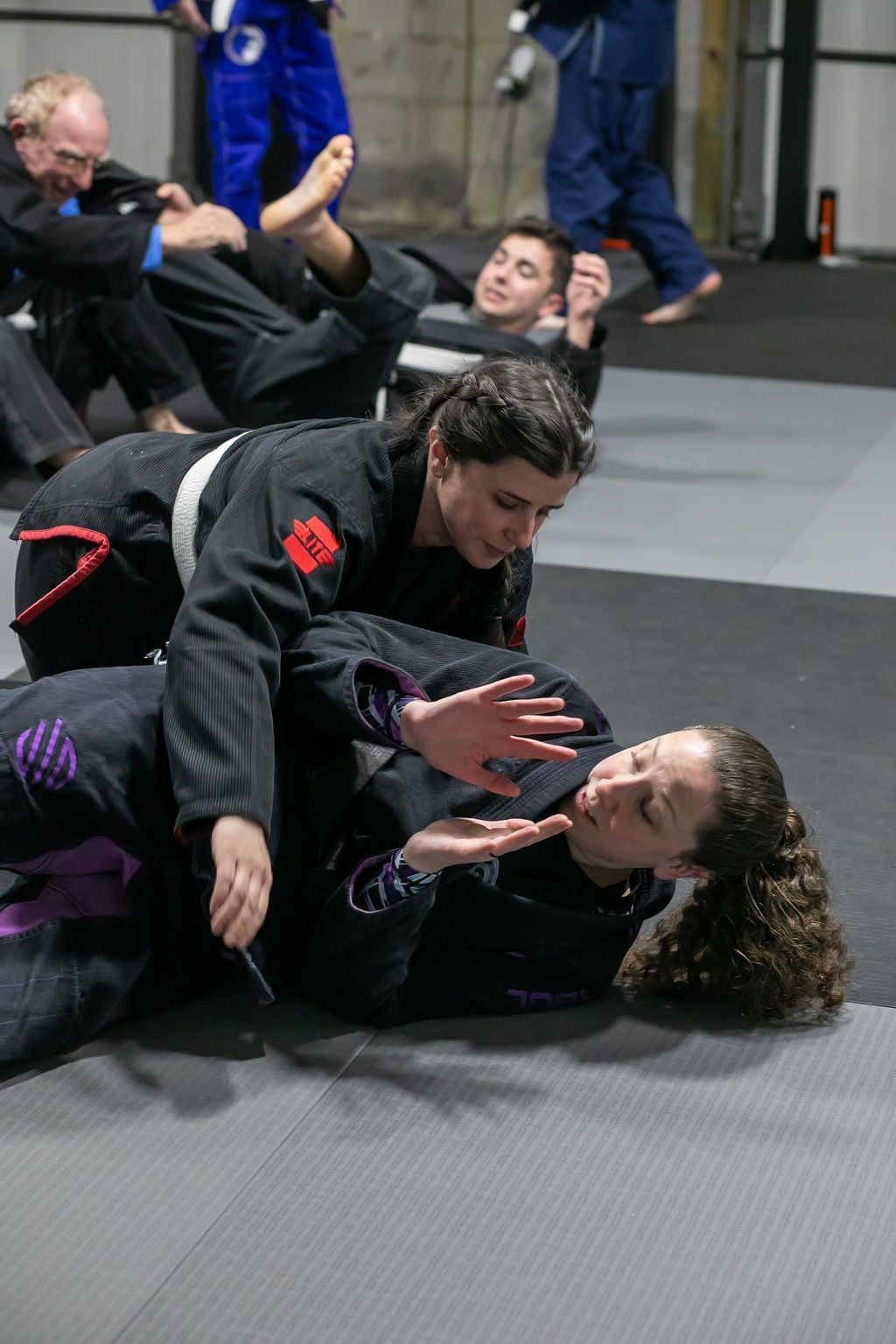 Two people practicing Brazilian jiu-jitsu on a padded gym mat, one pinning the other while others train behind them