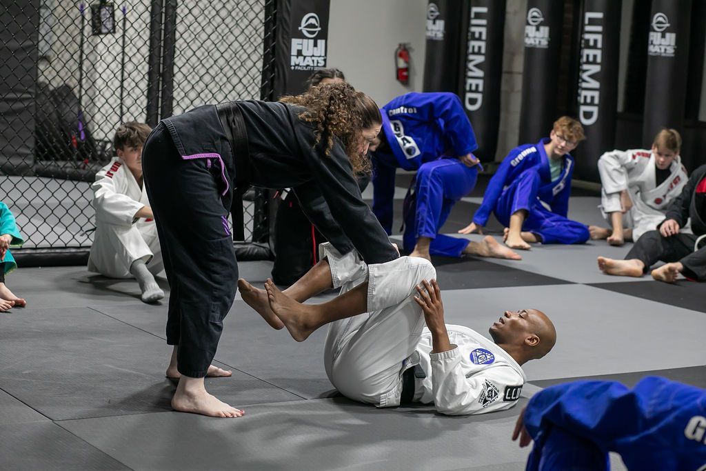 Two martial artists grappling on mats in a dojo, with two others blurred in the background.