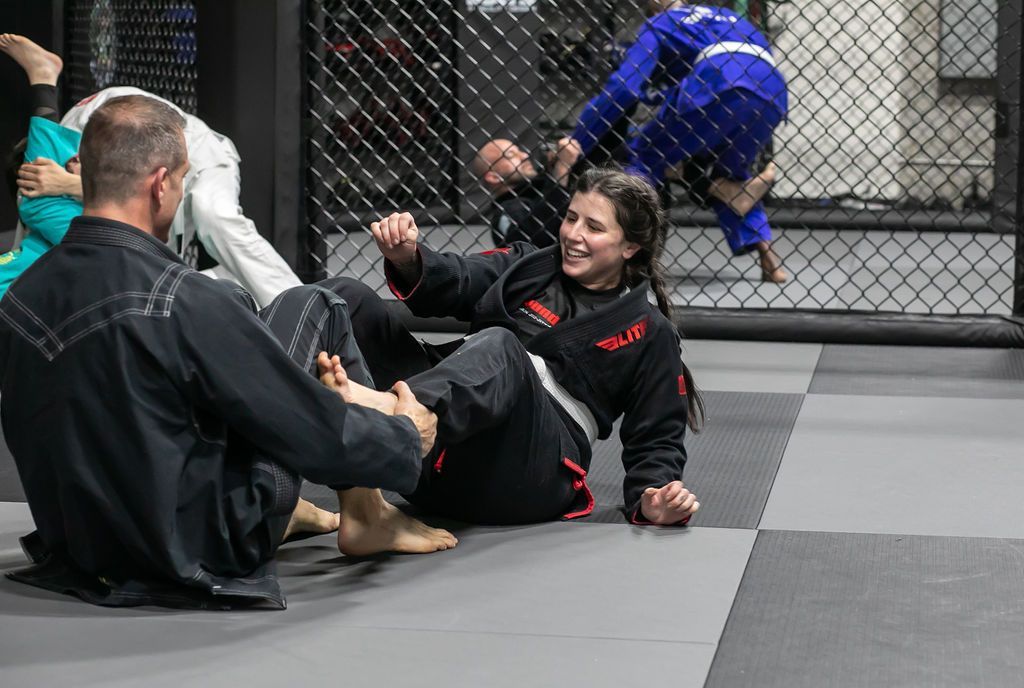 Martial arts class on mats, with two people practicing grappling near a chain-link fence.