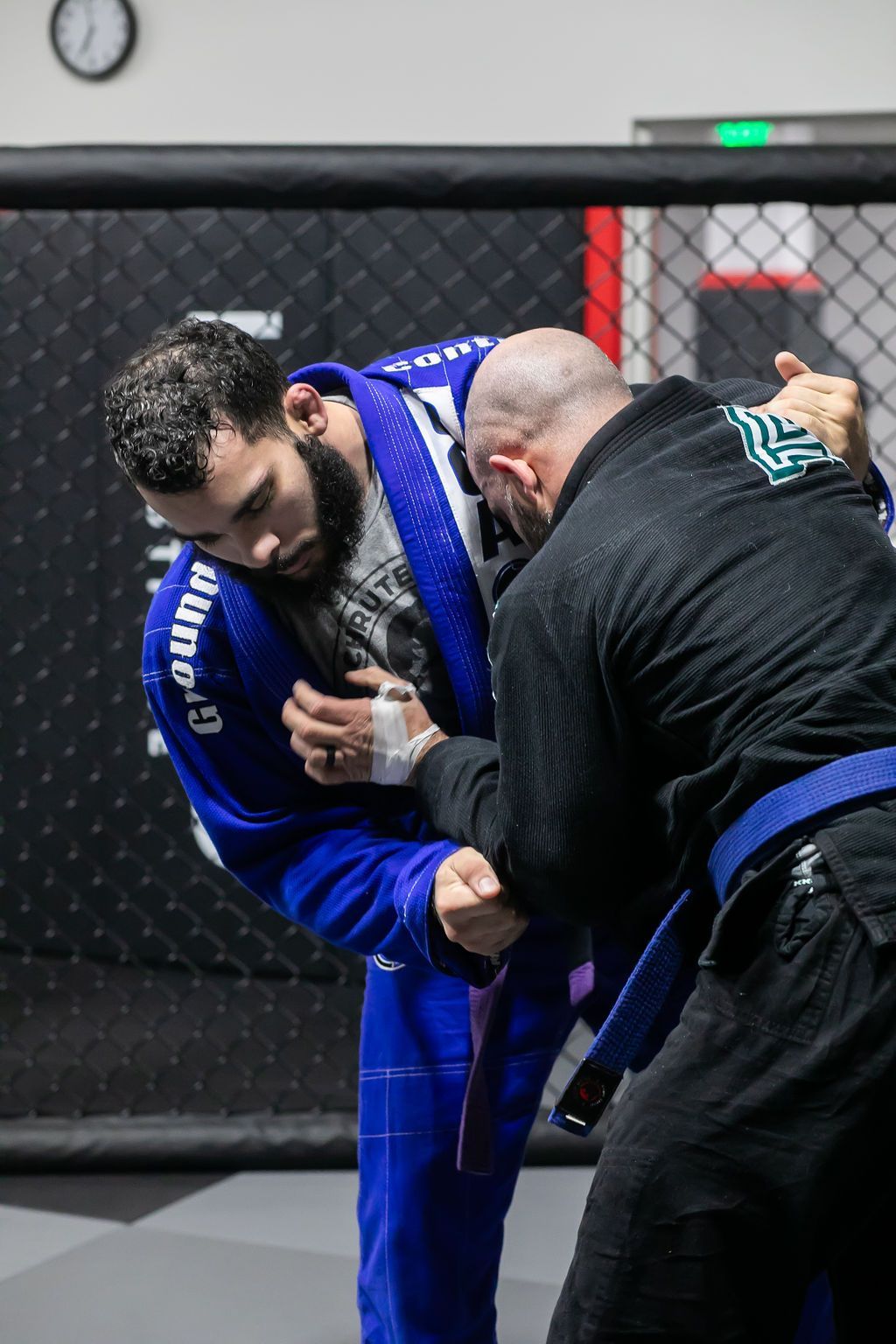 Two grapplers in blue and black gis practicing a clinch on a mat inside a fenced training area