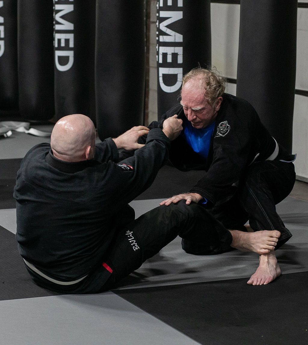 Two martial artists grappling on mats in a dojo, with two others blurred in the background.