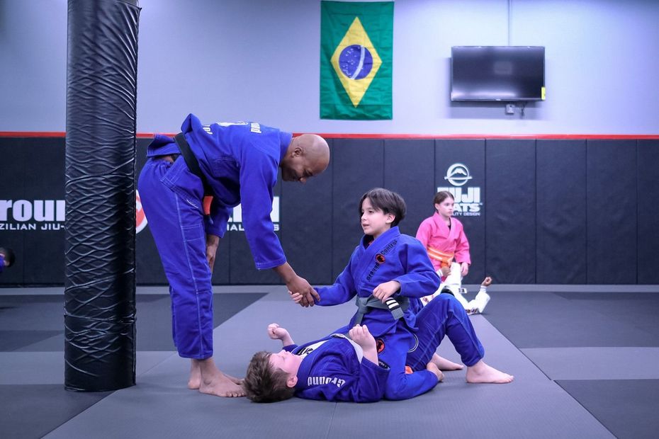 Instructor shaking hands with a student in blue gi, other child on the mat. Brazilian Jiu-Jitsu gym. Sparks Glencoe, MD.