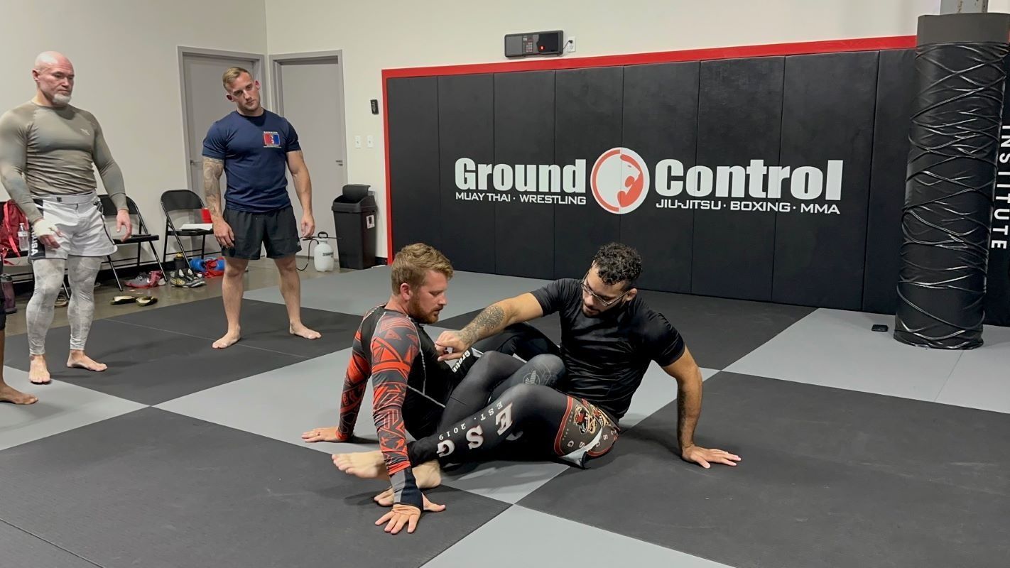 Two men in a grappling session; one seated, the other on top, in a gym with onlookers.