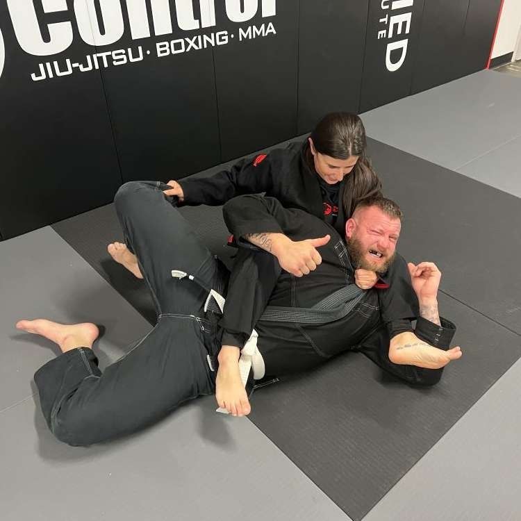 A young woman in black gi has a man in a black gi in a grappling hold on a mat. Man looks distressed.