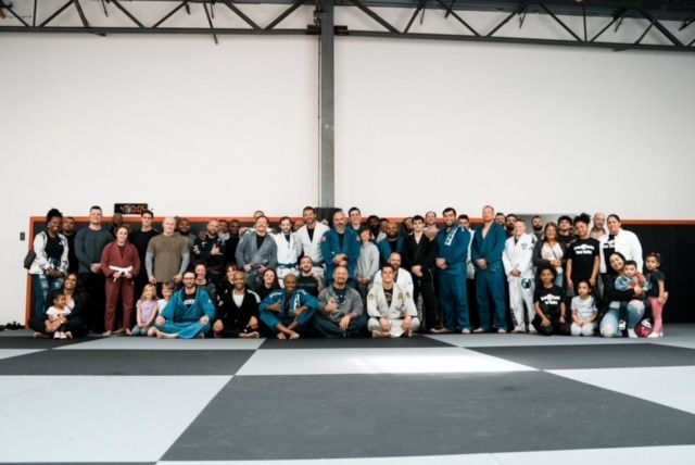 Group photo of many people in a martial arts gym, some wearing blue or white gis, smiling.