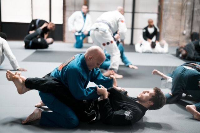 Two men in blue and black gis grappling on a mat, with other practitioners in the background.