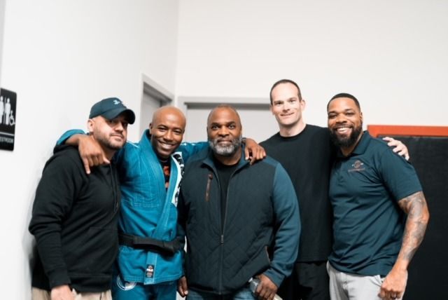 Five men smiling, posing together. One in a blue gi, others in casual wear. Inside a gym.