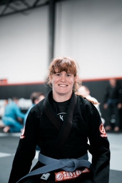 Woman in a black gi with a blue belt smiles in a martial arts training gym.