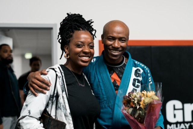 Smiling man in blue gi and woman with arm around him, holding flowers. Gym background.