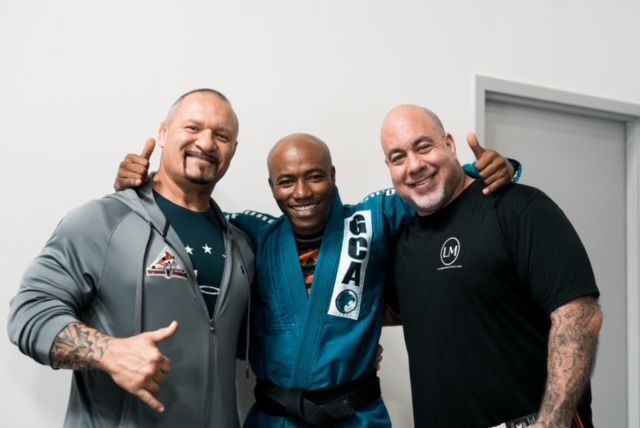 Three men smiling, posing together. Middle man in blue gi. Two men give hand gestures.