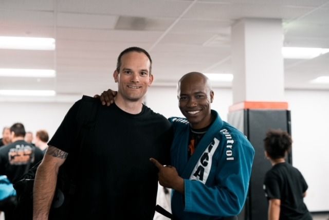 Two men in a gym smiling, one points. One wears a blue gi, the other a black t-shirt. White walls.