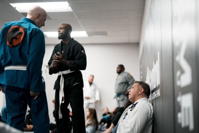 People in a jiu-jitsu gym: two men in gis talking, others training or resting.