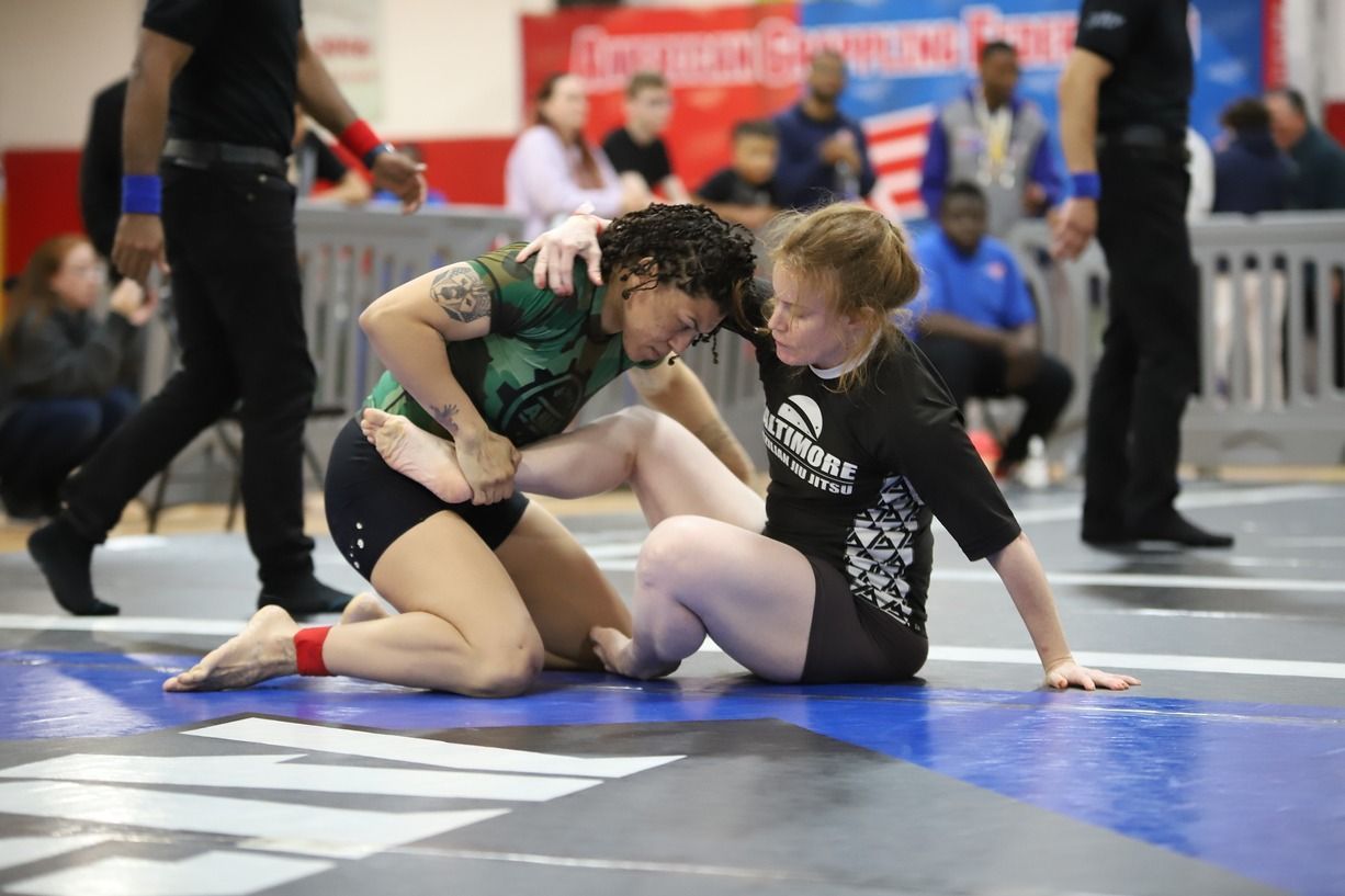 Two wrestlers grappling on a mat at an indoor competition, with spectators and barriers in the background. Sparks Glencoe, MD