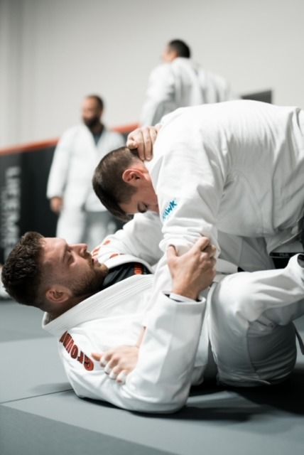 Two martial artists grappling on mats in a dojo, with two others blurred in the background.