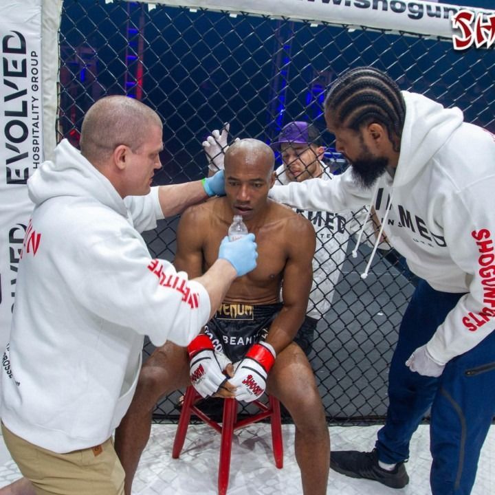 Two coaches tend to a fighter seated in a red chair inside an MMA cage between rounds