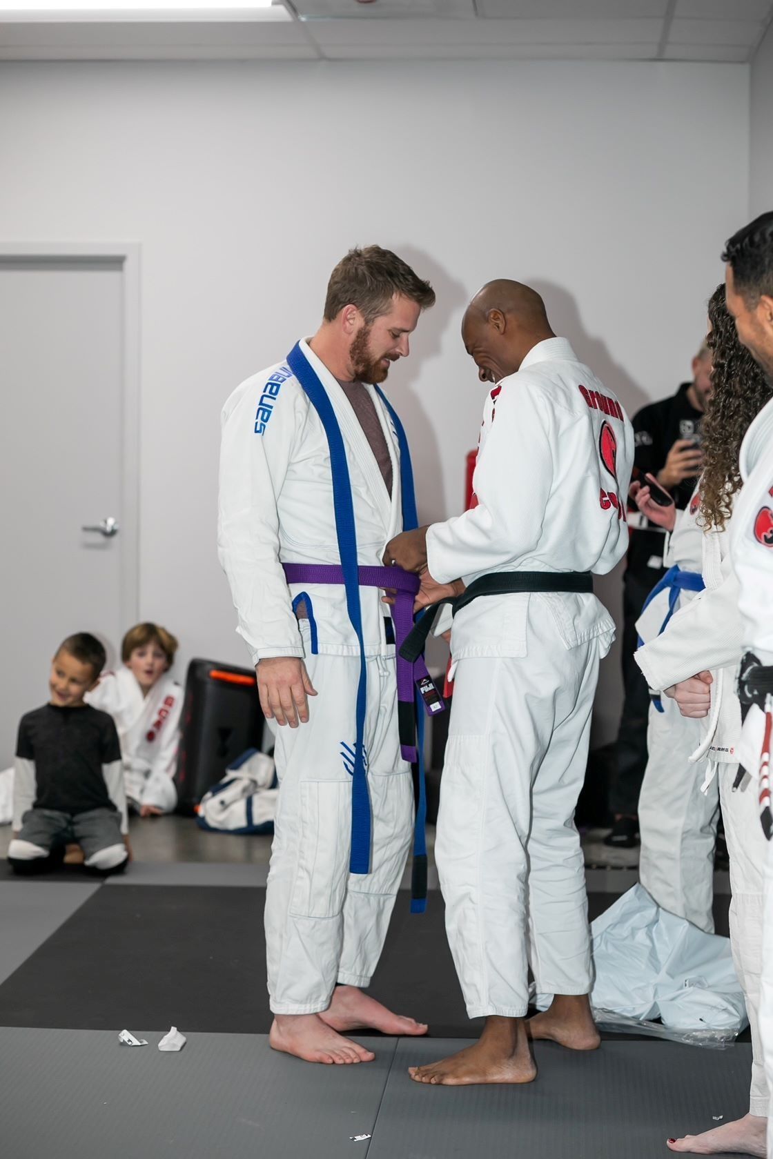 Man in white gi receiving a purple belt, indoors. Black belt adjusts the belt. Others watch.