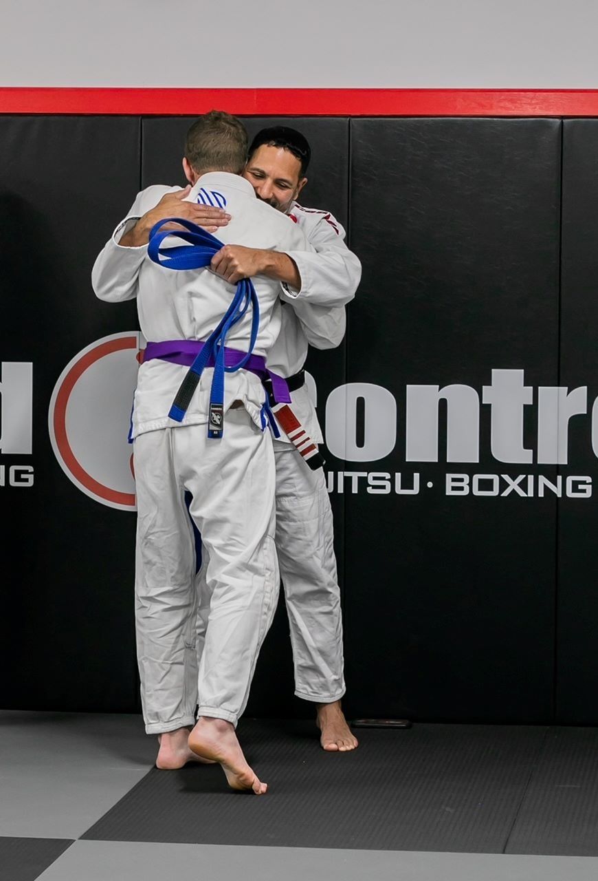 Two men in white gi uniforms embrace on a gym mat; one has a blue belt, the other a purple belt.